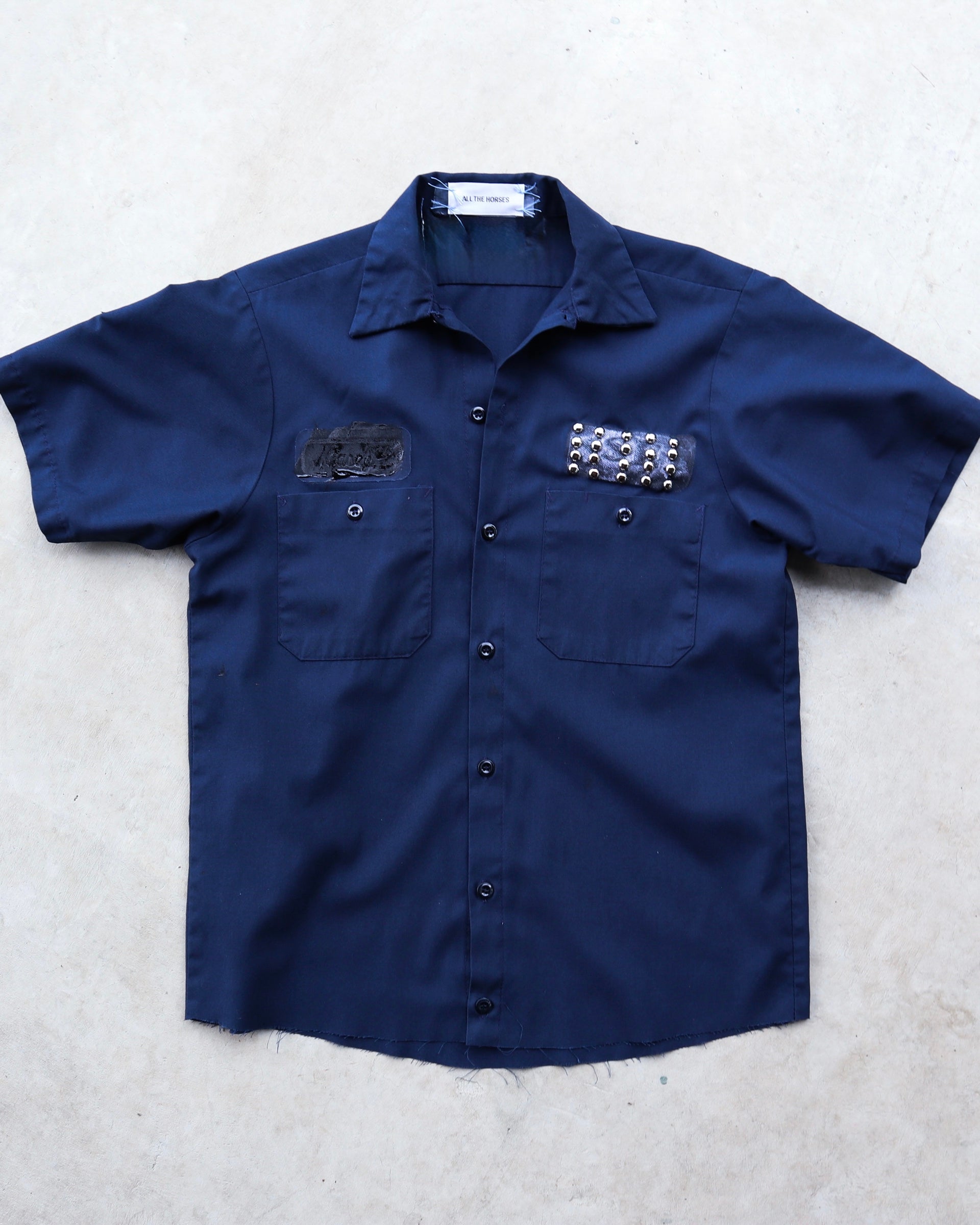 Navy one-of-one reworked mechanic shirt laid flat, with black chest patches, silver studs, and ATH label.